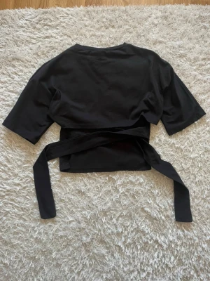 Svart croppad topp med knytband - Trendig svart croppad topp med korta ärmar och långa knytband i midjan som ger en snygg öppen rygg. Köpt från Nelly, använd fåtal gånger.