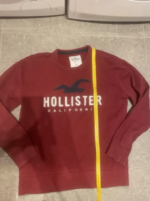 Vinröd sweatshirt från Hollister - Säljer en vinröd sweatshirt från Hollister med stort broderat logotyptryck och texten 'Hollister California' på bröstet. Tröjan har rund halsringning, långa ärmar och ribbade muddar. Perfekt för en avslappnad och stilren look. Stl M