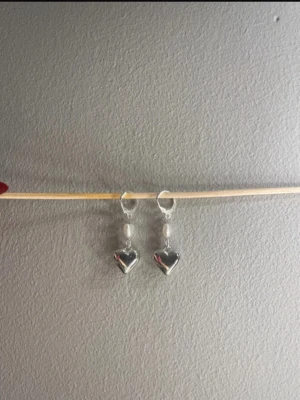 Silverörhängen med pärlhjärtan - Säljer ett par snygga örhängen i silverfärg med små pärlhjärtan och hängande hjärtberlocker. Örhängena har en modern och söt design som passar perfekt till dig som gillar detaljer. Materialet ser ut att vara metall och pärlimitation. Egengjorda 