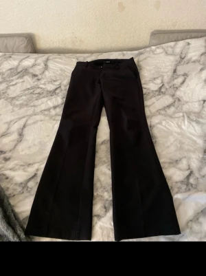 Svarta utsvängda kostymbyxor - Snygga svarta kostymbyxor med utsvängda ben och låg midja . Klassisk design med diskreta fickor och knappstängning fram. Perfekta för en stilren look och passar till många olika outfits. Materialet känns mjukt och följsamt.