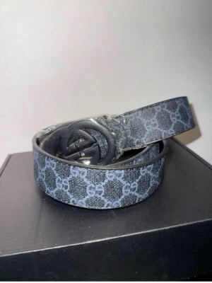 Gucci bälte - Snyggt svart och grått bälte från Gucci med klassiskt GG-mönster över hela bandet. Bältet har en rundad spänne i svart och är tillverkat i skinn med textilstruktur. Perfekt för att lyfta vilken outfit som helst.