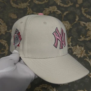 Beige New York Yankees keps - Snygg beige keps från New Era med rosa NY Yankees-logga framtill och broderad detalj på sidan. Klassisk böjd skärm och justerbar passform. Perfekt för dig som vill ha en stilren och trendig accessoar med sportig vibe.