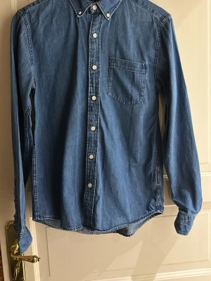 Blå jeansskjorta herr small Cubus - Snygg blå jeansskjorta från Cubus med klassisk krage, vita knappar och en bröstficka. 