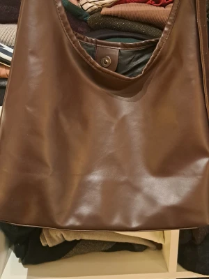 Brun axelremsväska i fejkläder - Stilren brun handväska i mjukt fejkläder med enkel axelrem och magnetknäppning. Väskan har en minimalistisk design och rymligt huvudfack, perfekt för dig som gillar klassiska accessoarer med modern touch.