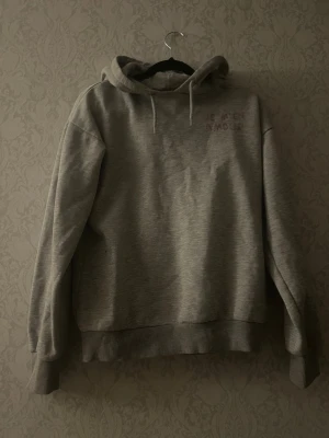 Grå hoodie med tryck 'JE M'EN AMOUR' - Säljer en grå hoodie med huva och dragsnören. På bröstet står det 'JE M'EN AMOUR' och på ryggen finns samma text i större tryck flera gånger tillsammans med ett hjärta. Hoodien har ribbade muddar vid ärmar och nederkant. Perfekt för en chill och avslappnad stil. Kanin finns i hemmet