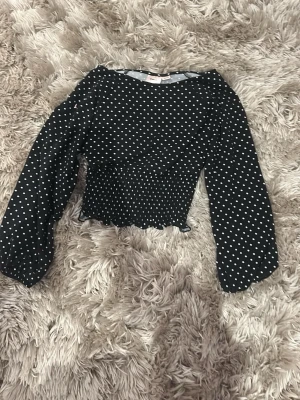 Svart prickig croppad topp - Säljer en svart croppad topp med vita prickar och långa ärmar. Toppen har en smockad midja och vågig kant nertill som ger en söt touch. Perfekt för dig som gillar trendiga och lekfulla plagg.