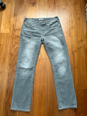 True religion jeans - Snygga grå jeans med raka ben och klassisk femficksdesign. Jeansen har en cool tvättad look och en unik ficka med lock bak. Perfekta för dig som gillar en avslappnad men ändå stilren vibe. Materialet är mjukt denim som sitter skönt hela dagen.