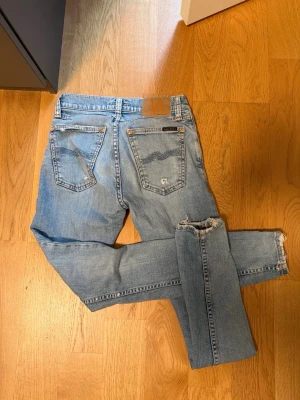 Ljusblå  jeans från Nudie - Säljer ett par ljusblå skinny jeans från Nudie med klassiska fem fickor och slitna detaljer vid benslut och bakfickor. Jeansen har en smal passform och är tillverkade i mjuk denim med Nudie-logga på bakfickan och läderpatch i midjan.