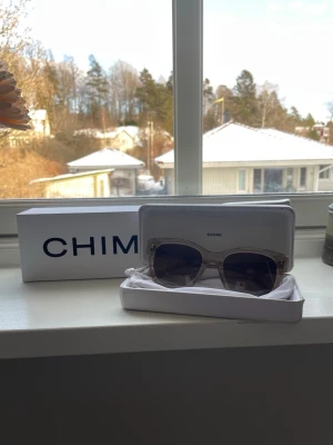 Transparenta solglasögon från Chimi - Säljer ett par stilrena solglasögon från Chimi med transparent båge och fyrkantig form. Glasen är tonade i brunt och ger en modern vibe. Perfekta för dig som vill ha en clean och trendig look.