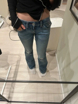 Blå low waist bootcut jeans - Säljer ett par blå low waist bootcut jeans med slitningar på knäna och snygga detaljer på bakfickorna, inklusive broderi och knapp. Jeansen har klassisk femficksmodell och är i ett mjukt denimtyg. Perfekta för dig som gillar en avslappnad men trendig look.