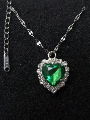 Halsband med hjärtformat grönt hänge - Snyggt halsband i silverfärgat rostfritt stål med ett hjärtformat grönt hänge omgivet av gnistrande klara stenar. Kedjan har ett elegant mönster och justerbar längd. Perfekt för dig som vill sticka ut med en färgklick och ett romantiskt smycke.