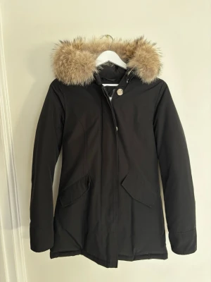 Svart parkas från Woolrich - Stilren svart parkas med huva från Woolrich. Jackan har två fickor framtill och stängs med dragkedja och knappar. 