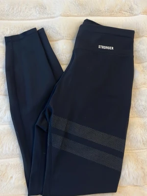 Svarta leggings från Stronger - Snygga svarta leggings från Stronger med hög midja och två diskreta randiga detaljer i mönster på ena benet. Tillverkade i stretchigt funktionsmaterial som passar perfekt till träning eller chill. Loggan sitter framtill i midjan. Nypris 700kr