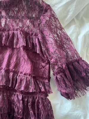 Vinröd spetsblus med volanger - Supersnygg vinröd blus i spets med flera lager volanger och hög hals. Blusen har trekvartsärm och är helt genombruten i spetsmaterial, vilket ger en romantisk och trendig look. Perfekt att styla med jeans eller kjol för en cool outfit.