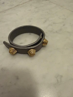 Balenciaga läderarmband med nitar - Snyggt svart läderarmband från Balenciaga med tre guldfärgade nitar i unik form. Armbandet har tryckknappsstängning och är perfekt för dig som vill ha en edgy accessoar med lyxig känsla. Märket är ingraverat på insidan. Strl S