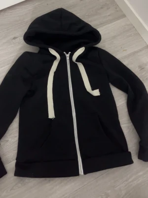 Svart hoodie med dragkedja - Svart hoodie med vit dragkedja och breda vita snören i huvan. Klassisk modell med fickor framtill och ribbade muddar vid ärmar och nederkant. Jag vet inte storleken på tröjan, men jag har XS och den skulle passat till högre storlekar😊