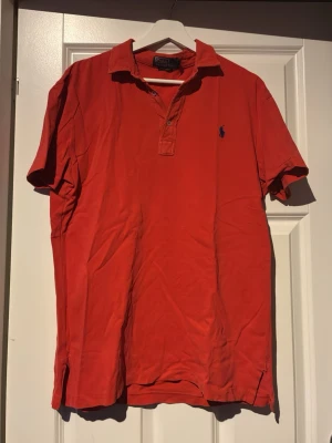Röd pikétröja från Polo Ralph Lauren - Klassisk röd pikétröja från Polo Ralph Lauren med blå broderad logga på bröstet. Tröjan har korta ärmar, krage och knappar vid halsen. Perfekt till jeans eller shorts för en avslappnad men snygg look.