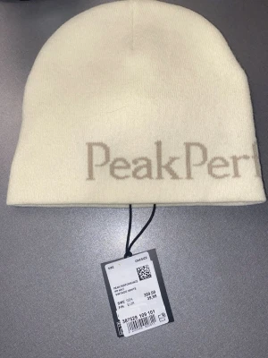 Vit mössa från Peak Performance - Snygg vit mössa från Peak Performance med diskret logga i beige framtill. Klassisk modell i mjukt och stretchigt material som sitter skönt på huvudet. Perfekt för kalla dagar och enkel att matcha med olika outfits.