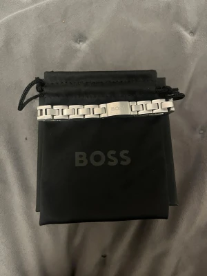 Silverfärgat armband från BOSS - Snyggt armband i silverfärgad metall från BOSS med stilren, länkad design och graverad logotyp på spännet. Kommer med originalpåse och ask, perfekt för dig som gillar minimalistiska accessoarer. Köpt för cirka en månad sedan och knappt använd för det va ej min stil. Köpt för 1300kr. Är i nyskick.