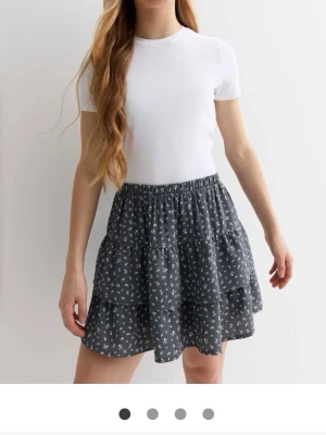 Mörkgrå blommig volangkjol - Supersöt mörkgrå kortkjol med små vita blommor och volangdetaljer. Kjolen har resår i midjan och är perfekt för vår och sommar. Lätt och luftig, passar till både t-shirt och stickad tröja.