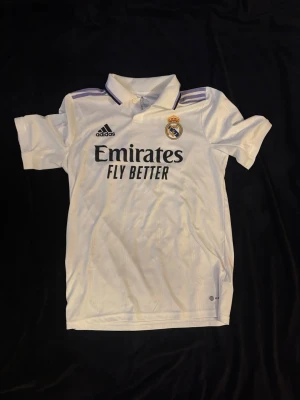 Real Madrid matchtröja Adidas vit ÄKTA - Säljer en vit Real Madrid matchtröja från Adidas med klubbmärke och sponsortryck på bröstet. Tröjan har korta ärmar, krage och lila detaljer på axlarna. Tillverkad i lätt och ventilerande material, perfekt för fotbollsträning eller match.