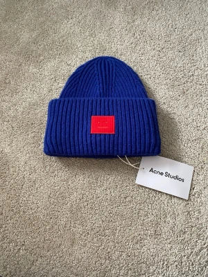 Blå ribbstickad mössa Acne Studios - Säljer en blå ribbstickad mössa från Acne Studios med uppvikt kant och det ikoniska röda Face-logotypen framtill. Mössan är i tjockt, mjukt material och har en modern, clean look som passar perfekt till streetwear och höst- eller vinteroutfits.