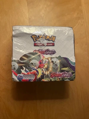 Pokémon Scarlet & Violet boosterbox - Säljer en Pokémon Trading Card Game boosterbox från Scarlet & Violet-serien. Förpackningen är plastad och har färgglad design med flera Pokémon på framsidan och sidan. Perfekt för samlare eller dig som vill bygga din kortsamling. OBS (ej äkta).