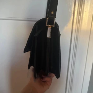 Svart axelremsväska i skinnimitation - Stilren svart handväska med axelrem och guldfärgade metalldetaljer. Väskan har en rektangulär form och slät yta, perfekt för dig som gillar minimalistisk design. Justerbar rem gör den enkel att bära över axeln. Passar till många olika outfits.