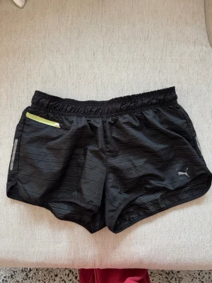 Svarta träningsshorts från Puma - Säljer ett par svarta träningsshorts från Puma med diskreta ränder och elastisk midja. Shortsen har en liten ficka med dragkedja i neonfärg och är tillverkade i lätt polyester, perfekta för löpning eller gymmet.
