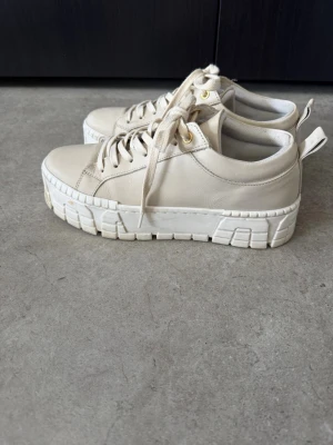 Beige chunky sneakers med platåsula - Stilrena beige sneakers med kraftig vit platåsula och snörning. Skorna har en modern chunky design och är tillverkade i ett slätt material med detaljer i guld vid snörhålen. Perfekta för dig som gillar trendiga och bekväma skor.