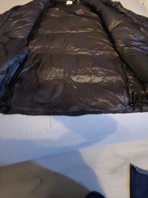 Svart dunjacka med huva - Säljer en svart dunjacka med huva och dragkedja samt tryckknappar framtill. Jackan har två stora fickor med dragkedja och är quiltad för extra värme. Perfekt för kalla vinterdagar och har en klassisk, stilren look.