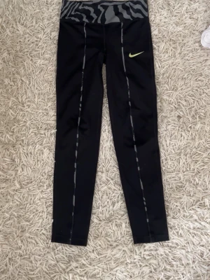 Nike svarta leggings med mönstrad midja - Svarta leggings från Nike med grått zebramönster i midjan och neongul logga på låret. Tight passform och långa ben, perfekta för träning eller chill. Snygga kontrastsömmar längs benen. Materialet är stretchigt och följsamt.