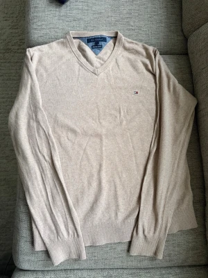Beige V-ringad tröja Tommy Hilfiger - Snygg beige stickad tröja från Tommy Hilfiger i Pima Cotton Cashmere. V-ringad modell med diskret logga på bröstet och ribbade muddar vid ärmslut och nederkant. Perfekt för lager på lager och stilrena outfits.