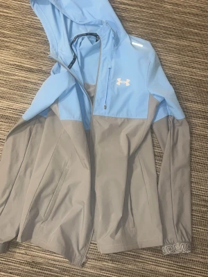 Blå och grå vindjacka från Under Armour - Snygg vindjacka från Under Armour i ljusblått och grått. Jackan har huva, dragkedja och logotyp på bröstet. Materialet är lätt och funktionellt, perfekt för att skydda mot vind. Ärmslut med tryckt detalj och modern passform.