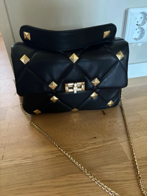 Svart Valentino handväska med gulddetaljer - Snygg svart handväska från Valentino Garavani i quiltat läder med guldiga nitar och kedja. Väskan har ett elegant spänne framtill och bred axelrem med logga. Perfekt accessoar för att lyfta din outfit.