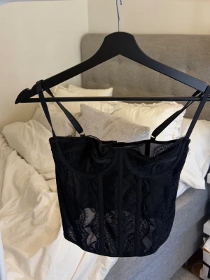 Svart spetskorsett med justerbara band - Snygg svart korsettlinne med spetsdetaljer och justerbara axelband. Korsetten har en markerad midja och hakknäppning i ryggen. Materialet är en mix av spets och tyg som ger en elegant look. Perfekt för att skapa en feminin siluett. (oanvänd)