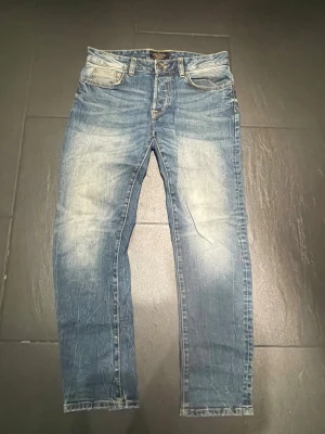 Blå jeans - Säljer ett par blå slim/raka jeans från jack and jones med snyggt slitna detaljer och klassisk femficksmodell. Jeansen har en ljus tvätt med tydliga slitningar på lår och knän, samt gylf med knappstängning. Perfekta för en avslappnad och trendig look. Jeansen är lagade i skrevet men inget som syns eller känns när man har på sig de:)