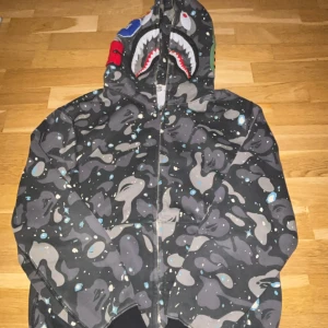 BAPE Shark Hoodie med camomönster - Säljer en BAPE Shark Hoodie i storlek L med grått och svart camomönster, blå och vita detaljer samt den ikoniska hajdesignen på huvan. Tröjan har dragkedja framtill och ribbade muddar. Perfekt för dig som vill sticka ut med streetwear.