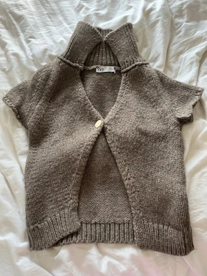 Beige stickad kofta från Zara - Kortärmad stickad kofta från Zara i beige nyans. Koftan har en hög krage som man kan vika ner, en knapp framtill och ribbstickade detaljer. Perfekt för lager-på-lager och en chill look. Skriv gärna prisförslag 💕