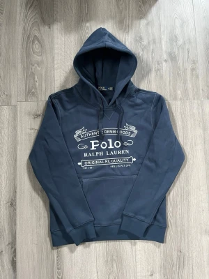 Mörkblå hoodie från Polo Ralph Lauren - Snygg mörkblå hoodie från Polo Ralph Lauren med stor vit logga och text på bröstet. Klassisk känguruficka framtill och justerbar dragsko i huvan. Tillverkad i mjukt bomullsmaterial som är skönt att bära. Perfekt för en avslappnad och trendig look.