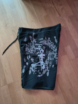 Svarta billabong shorts med tryck - Säljer ett par svarta shorts från billabong med stort vitt och grått grafiskt tryck på sidan. Shortsen har snörning i midjan och en ficka bak. Perfekta för sommaren och streetwear-stil.
