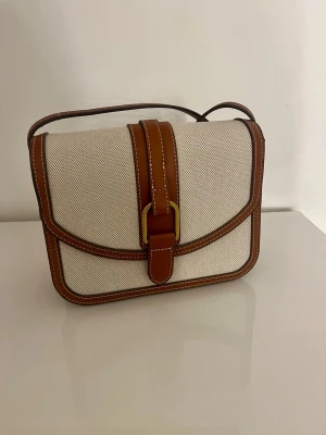 Väska  - Stilren axelväska i beige canvas med bruna läderdetaljer och vit söm. Väskan har en rektangulär form, justerbar axelrem och guldfärgat spänne framtill. Perfekt för dig som gillar klassisk och tidlös design. Rafiaeffekt. Bomullsblandningstyg. Kontrastdetalj. Långt justerbart handtag. Stängs med lock med magnetknapp. Innerficka, innerfoder och mått: 22x18x7 