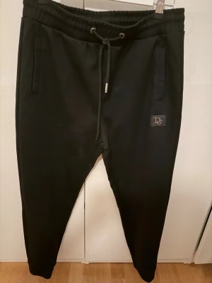 Svarta mjukisbyxor från DR - Svarta mjukisbyxor från DF med dragsko i midjan och två sidofickor. Byxorna har en slim fit-modell och en liten logotyp på vänster ben. Perfekta för en avslappnad stil och tillverkade i mjukt material.