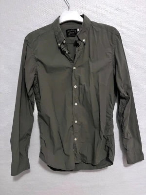 Olivgrön skjorta från Morris, S - Olivgrön skjorta från Morris med klassisk krage och vita knappar framtill. Långärmad modell i 100% mjuk bomull, med broderad logga på bröstet. Snygg och stilren passform som funkar till många olika looks. Perfekt för dig som gillar enkel och clean stil.