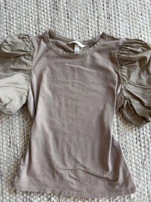 Beige topp med puffärmar från H&M -  Beige topp från H&M med korta, voluminösa puffärmar och rund halsringning. Tillverkad i mjuk bomullsmix som känns skön mot huden. Perfekt för dig som vill ha en stilren men ändå unik look.