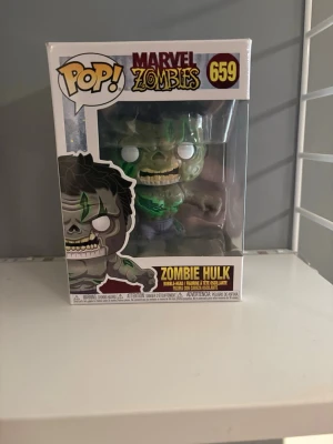 Funko Pop Marvel Zombie Hulk 659 - Cool samlarfigur från Funko Pop, Marvel Zombies-serien. Det är Zombie Hulk, nummer 659, i grön och grå färg med vita ögon och detaljerad zombie-look. Figuren kommer i originalförpackning med fönster och tryck.