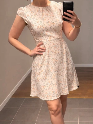 Blommig kortklänning i ljusa toner - Söt kortklänning med ljust blommigt mönster i vitt, rosa och grönt. Klänningen har rund halsringning, korta ärmar och markerad midja som ger en feminin siluett. Perfekt för vår och sommar. Materialet känns lätt och luftigt.