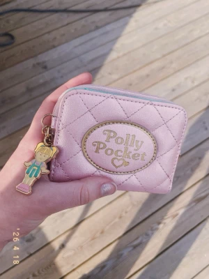Rosa Polly Pocket plånbok med nyckelring - Supersöt ljusrosa plånbok från Polly Pocket med quiltat mönster och gulddetaljer. Framsidan har ett broderat Polly Pocket-märke och dragkedjan har en charmig Polly Pocket-figur som nyckelring. Baksidan har ett fack med plastfönster för kort eller ID. 💕