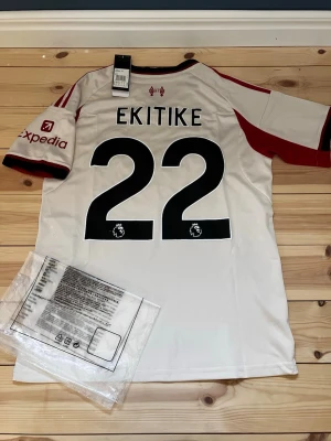 Liverpool bortatröja Ekitike #22 - Snygg Liverpool FC bortatröja från Adidas med Ekitike och nummer 22 på ryggen. Tröjan är vit med röda detaljer, klubbmärke och sponsorloggor på bröstet. Tillverkad i lätt och ventilerande material med Aeroready-teknologi. Perfekt för fotbollsträning eller match. Väldigt bra kvalitet