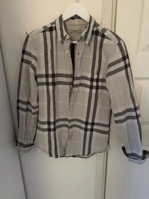Burberry Brit skjorta - Säljer en klassisk skjorta från Burberry Brit i vitt och grått. Skjortan har långa ärmar, knappar framtill och den ikoniska Burberry-rutan. Perfekt för dig som gillar stilrena och tidlösa plagg. Storlek S. Bra skick
